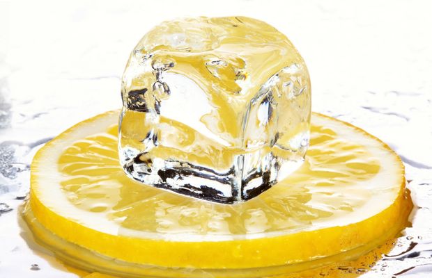 A slice of lemon under an ice cube on a wet white surface